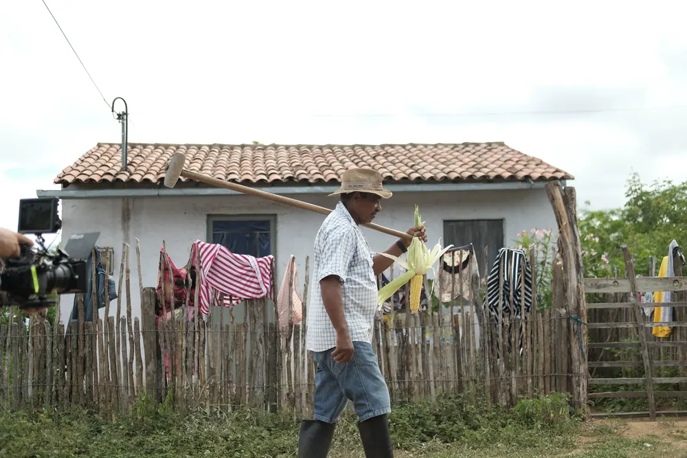 Agricultor caminha com a enxada no ombro diante da casa.