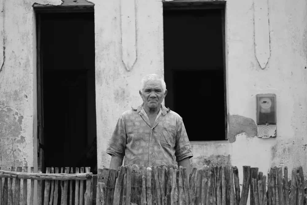 Senhor à janela de uma casa antiga, atrás da cerca de varas.